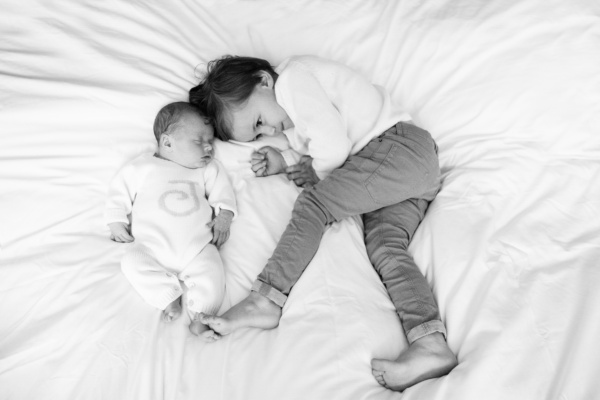 A newborn and a sibling snuggle together on a bed.