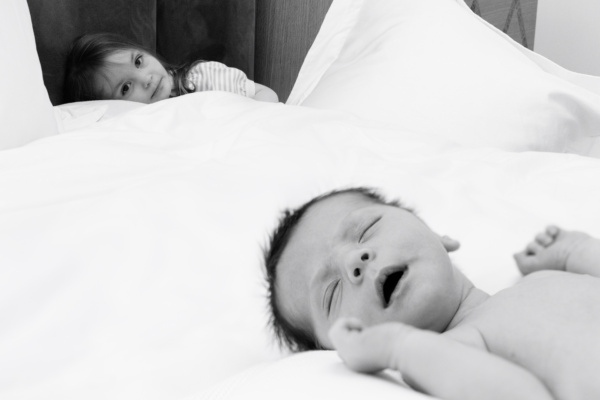 A newborn sleeps on their parents' bed while their big sister watches.