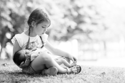 An older sister cradles their newborn sibling while sitting cross-legged on the grass.