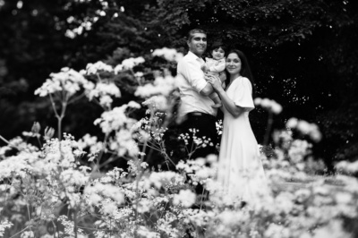 Parents and her baby stand behind flowers in a park.