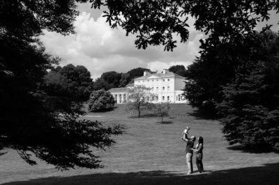 Parents hold up their baby at Kenwood House, London, framed by trees.