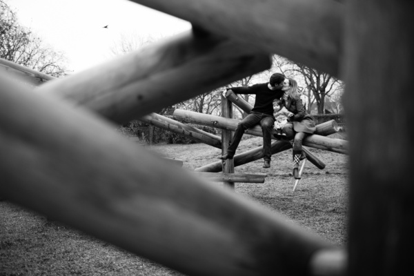 Parents kiss in a playground, framed by wooden playground equipment.