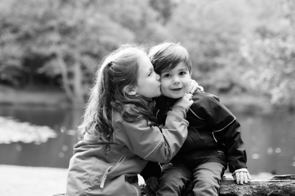 A sister kisses her brother on the cheek with a lake in the background.