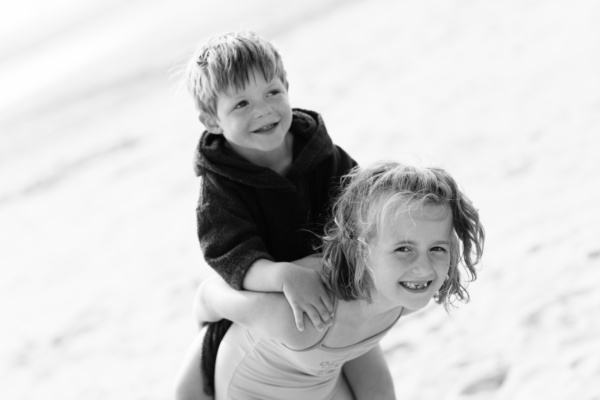 A sister piggybacks her brother across a beach.