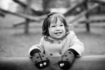 A smiling girl wearing ladybug gloves leans on a fence.