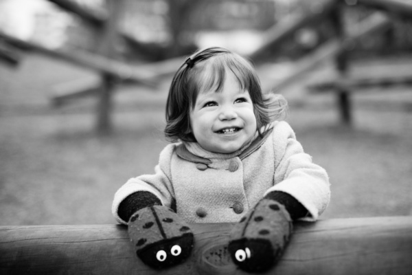 A smiling girl wearing ladybug gloves leans on a fence.