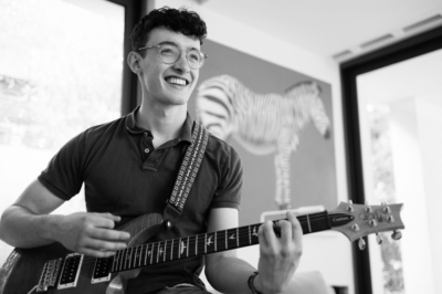 A smiling teenager plays the guitar with a zebra in the background.