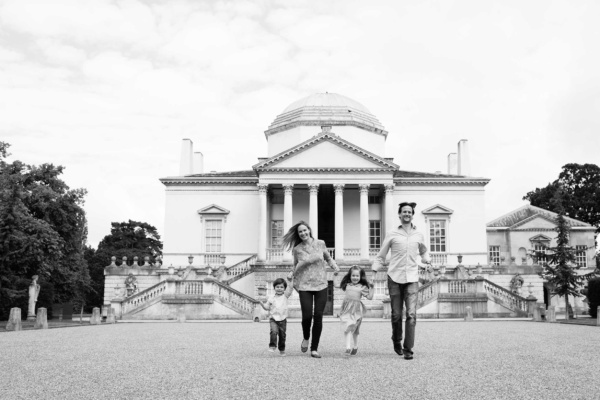 A family of mother, father, and two young children run towards the camera holding hands and smiling with Chiswick House in the background.