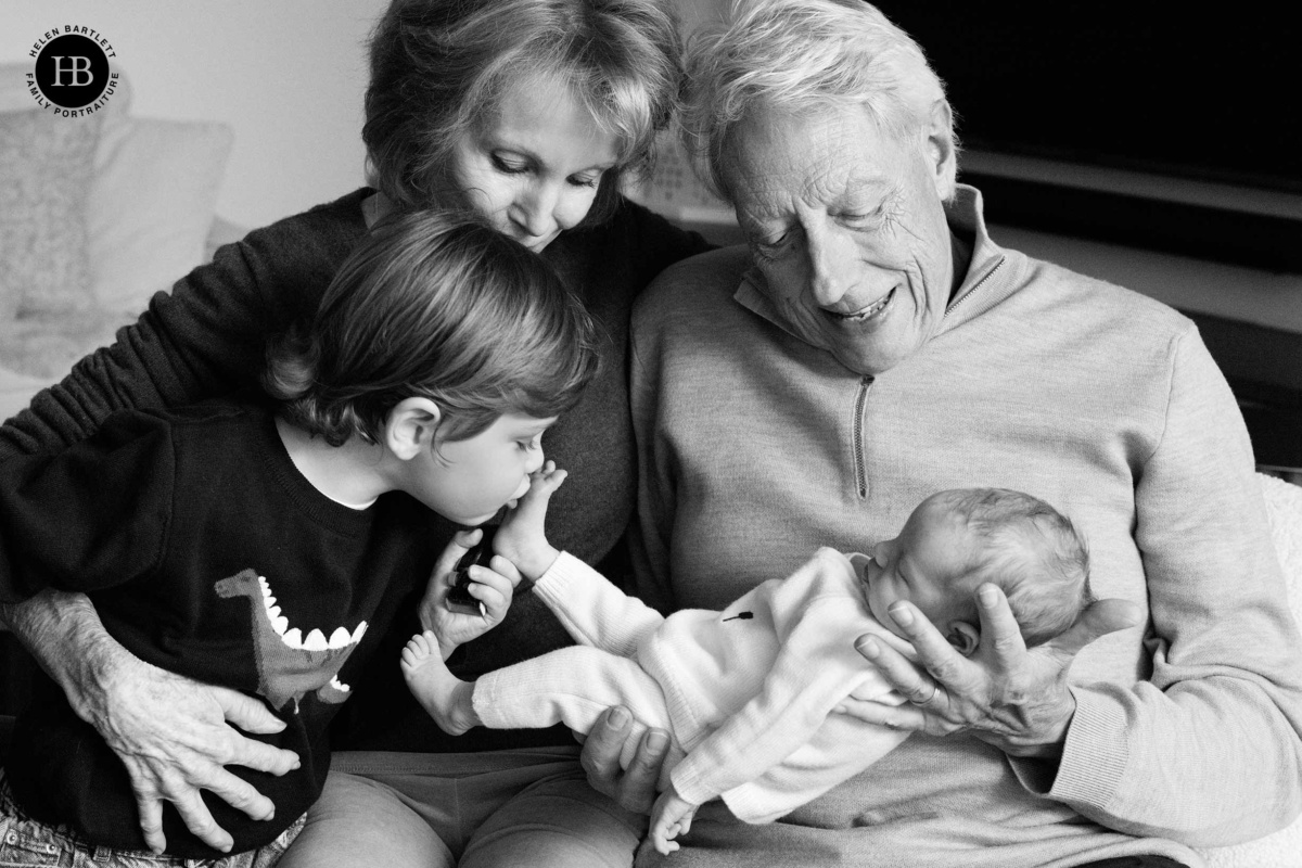 Young boy kissing the foot of his newborn baby sister while grandparents look on