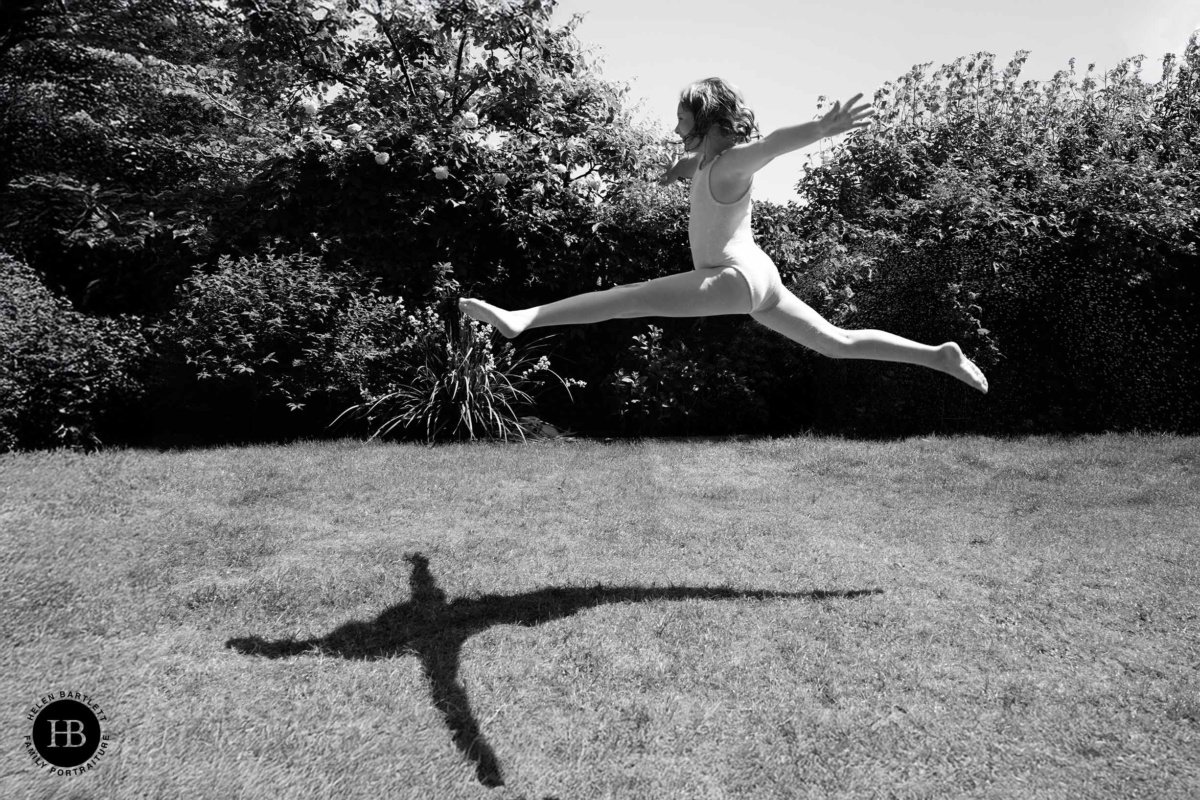 Girl leaping in mid-air in the garden, her shadow forming a cross shape on the lawn below