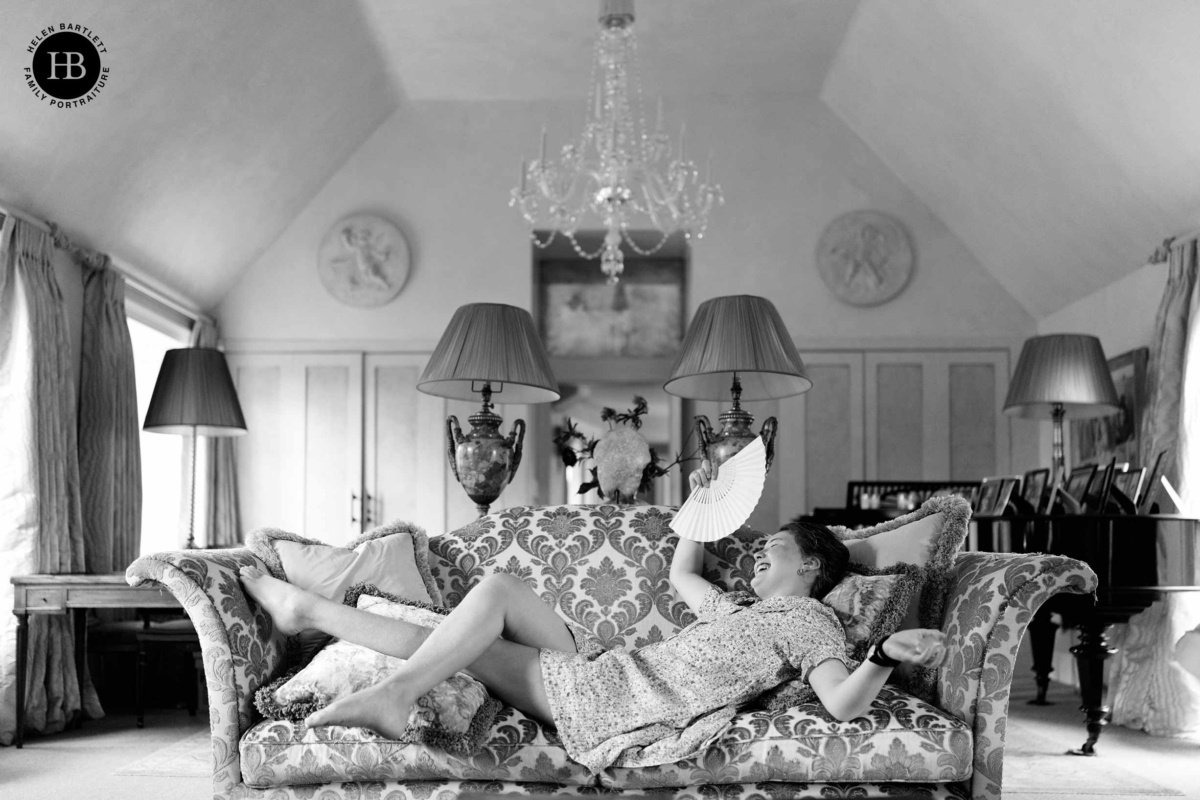 Teenage girl lying back on a sofa beneath a chandelier, laughing and waving a paper fan