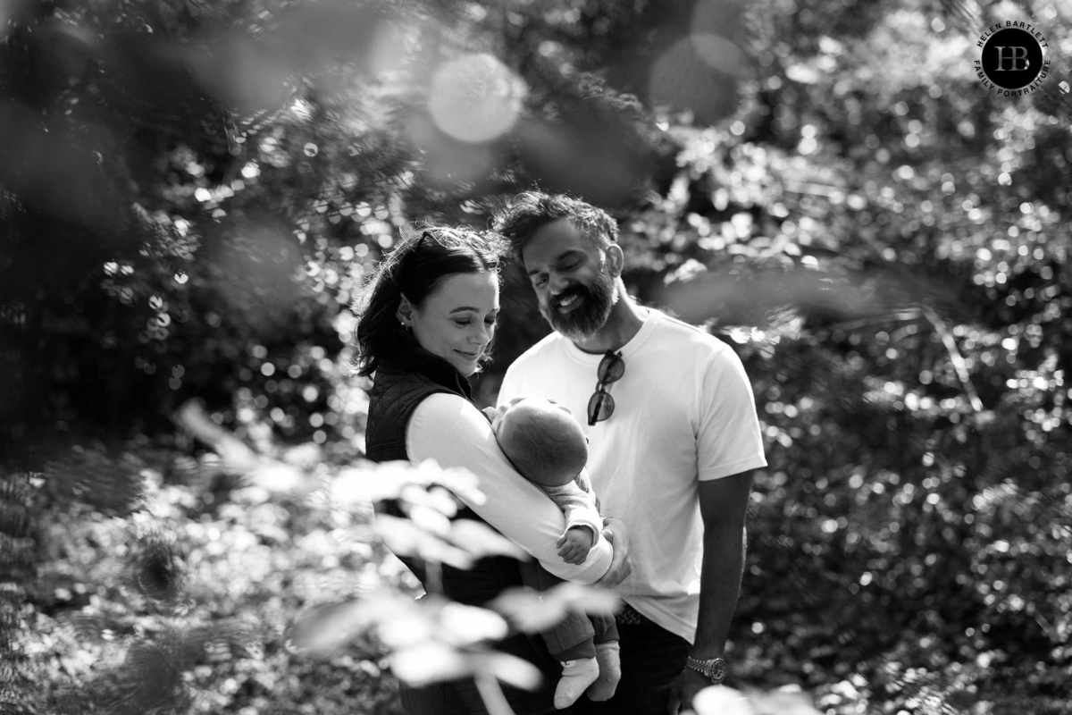 Mother and father smiling down at their baby, framed by dappled woodland leaves