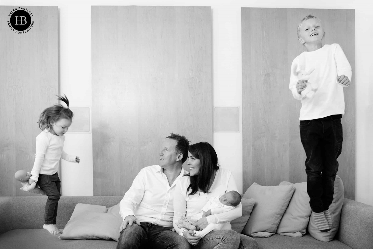 family photo with mum and dad holding newborn baby and older siblings jumping on sofa