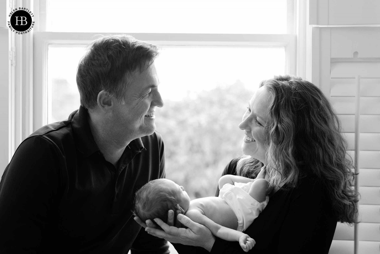 Family photo of mum, dad and newborn in font of window. Parents laughing and happy