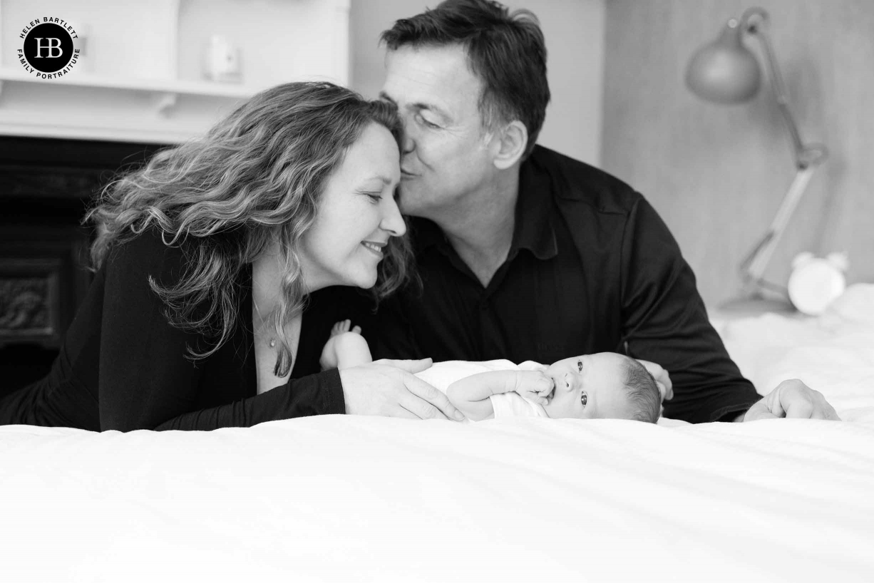 dad kisses mum while baby lies on bed in newborn baby family photoshoot Dulwich