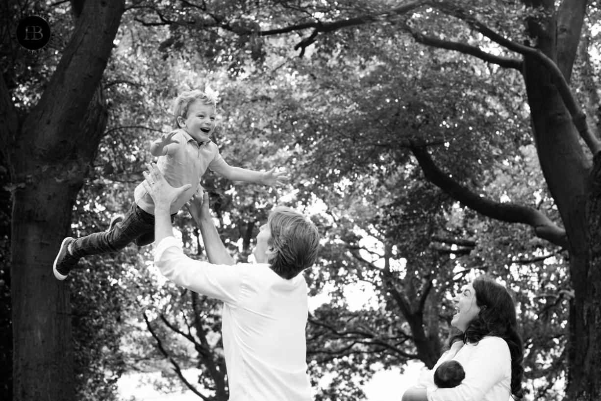 Dad throws son in the air while mum and newborn daughter look on. Fun family photo on Hampstead Heath