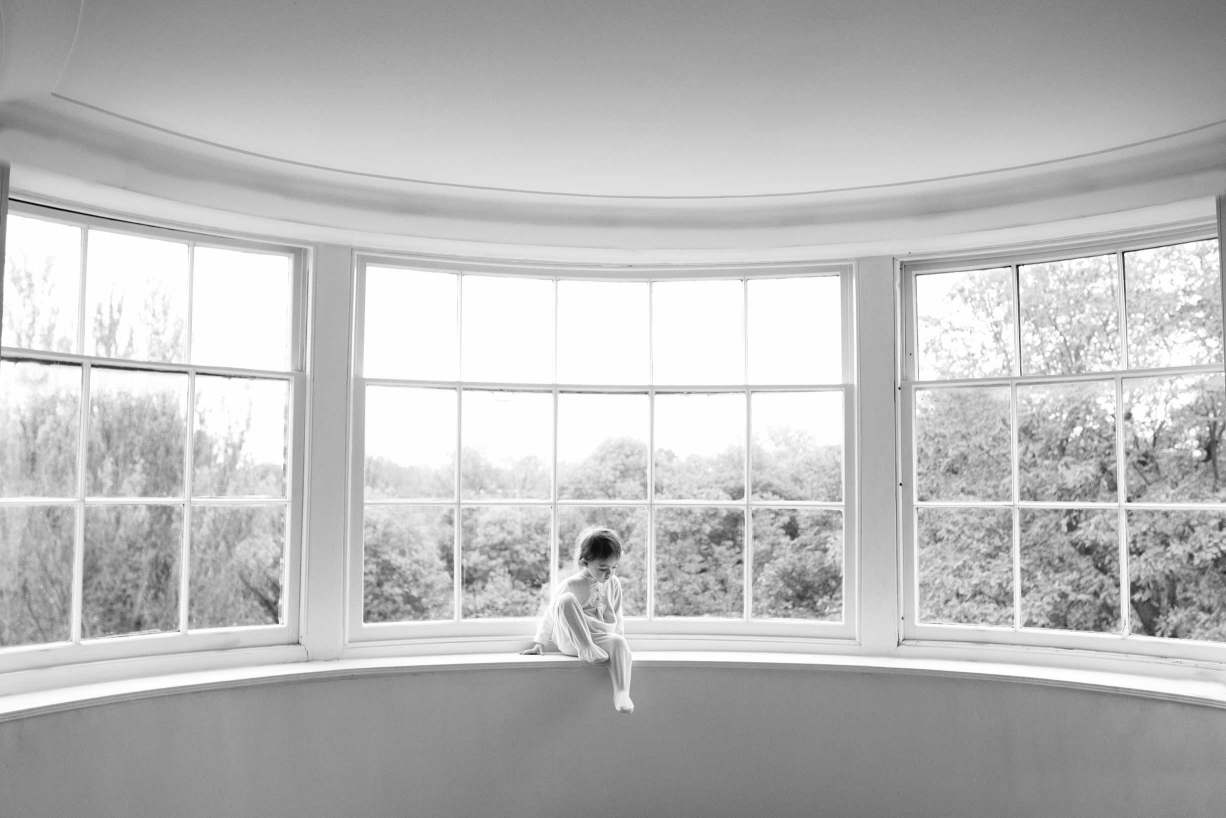 A tiny child sits in a window for this timeless portrait in black and white