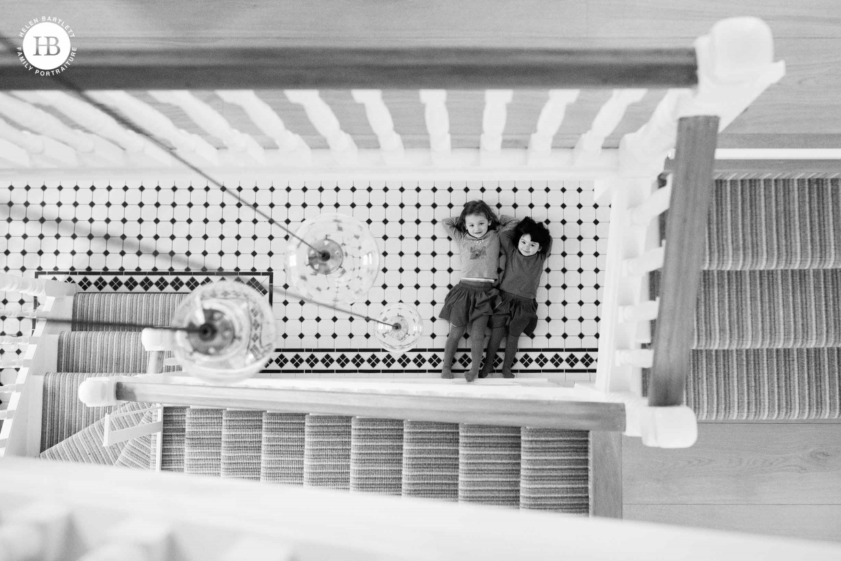 Family photography at home: two girls lie on the hall floor looking up at the photographer. The image is taken from three floors up and the stairs spiral around showing the interior of their London home