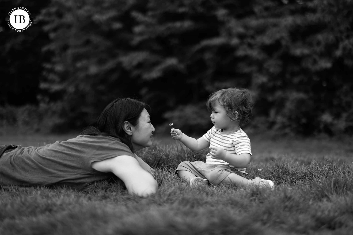 One year old baby offers a buttercup to mother who is lying on the grass with him. Black and white image with shallow depth of field.