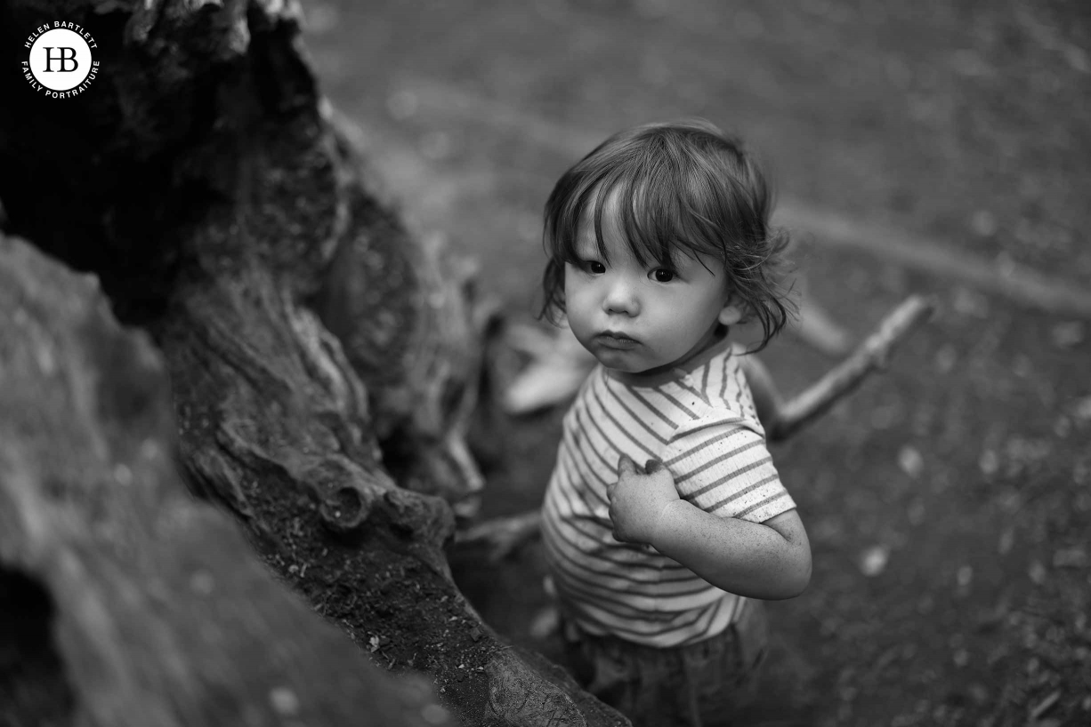 Little boy looks up and points to himself. He’s playing with mud and sticks by a fallen tree.