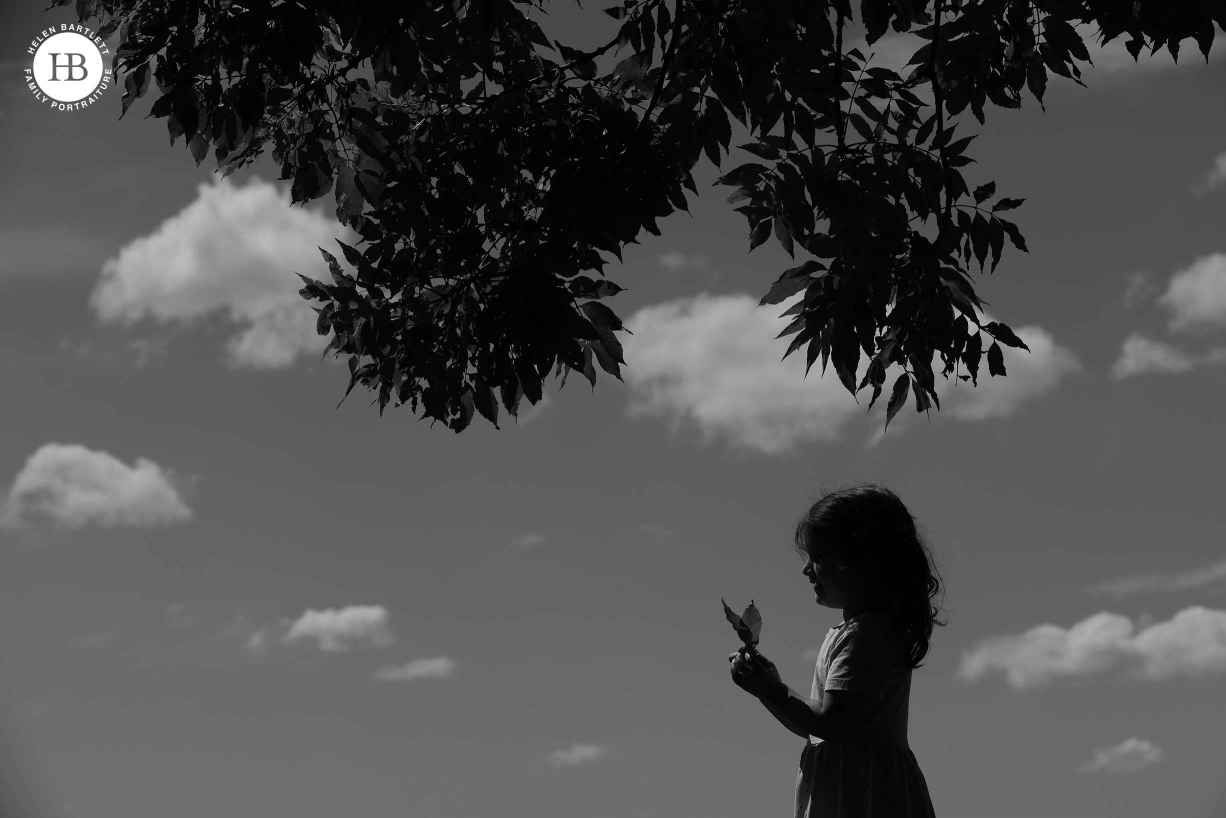 Silhouette of little girl holding two leaves and framed buy the tree above. White clouds in the background show beautiful hot day.