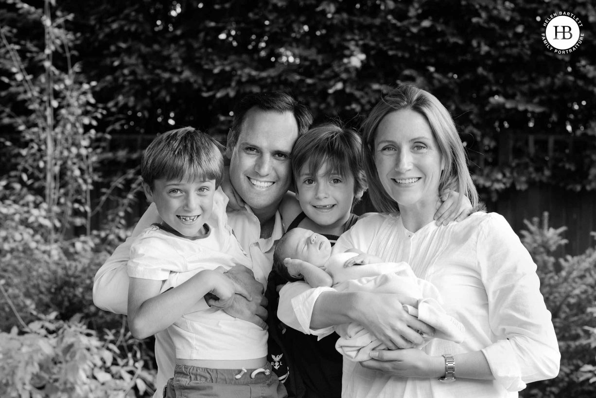 family of five hold newborn baby in outdoor family portrait. Everyone is laughing.