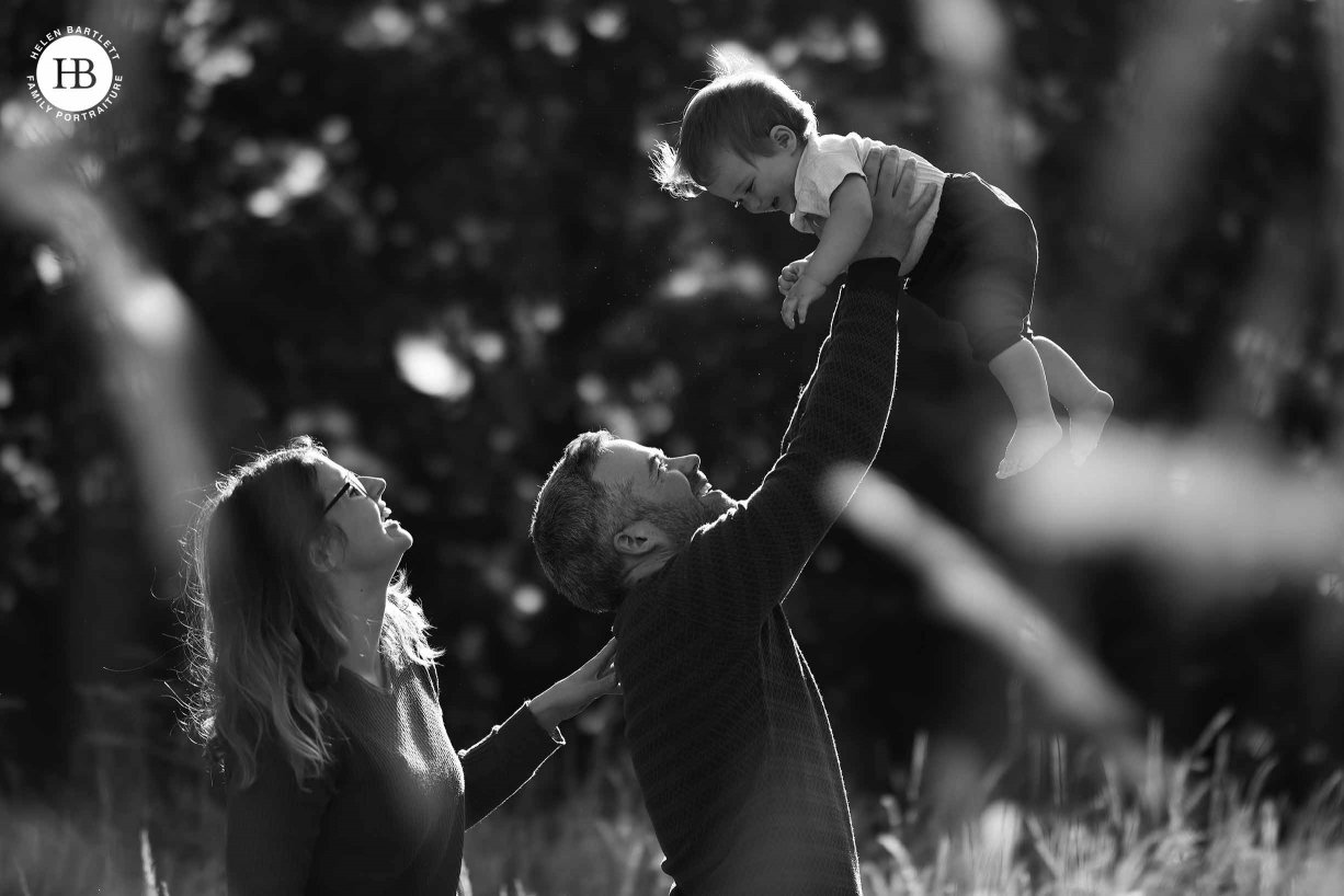 Dad, mum and baby shot in profile as dad lifts baby above his head. Backlit for beautiful effect and shot through long grass for foreground detail. Image used as an illustration for beautiful files from Canon EOS R5