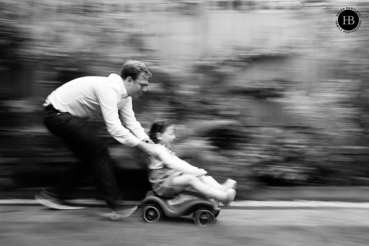 Dad pushes daughter on toy car in Hampstead Garden. Panning technique used to show speed in the photograph.