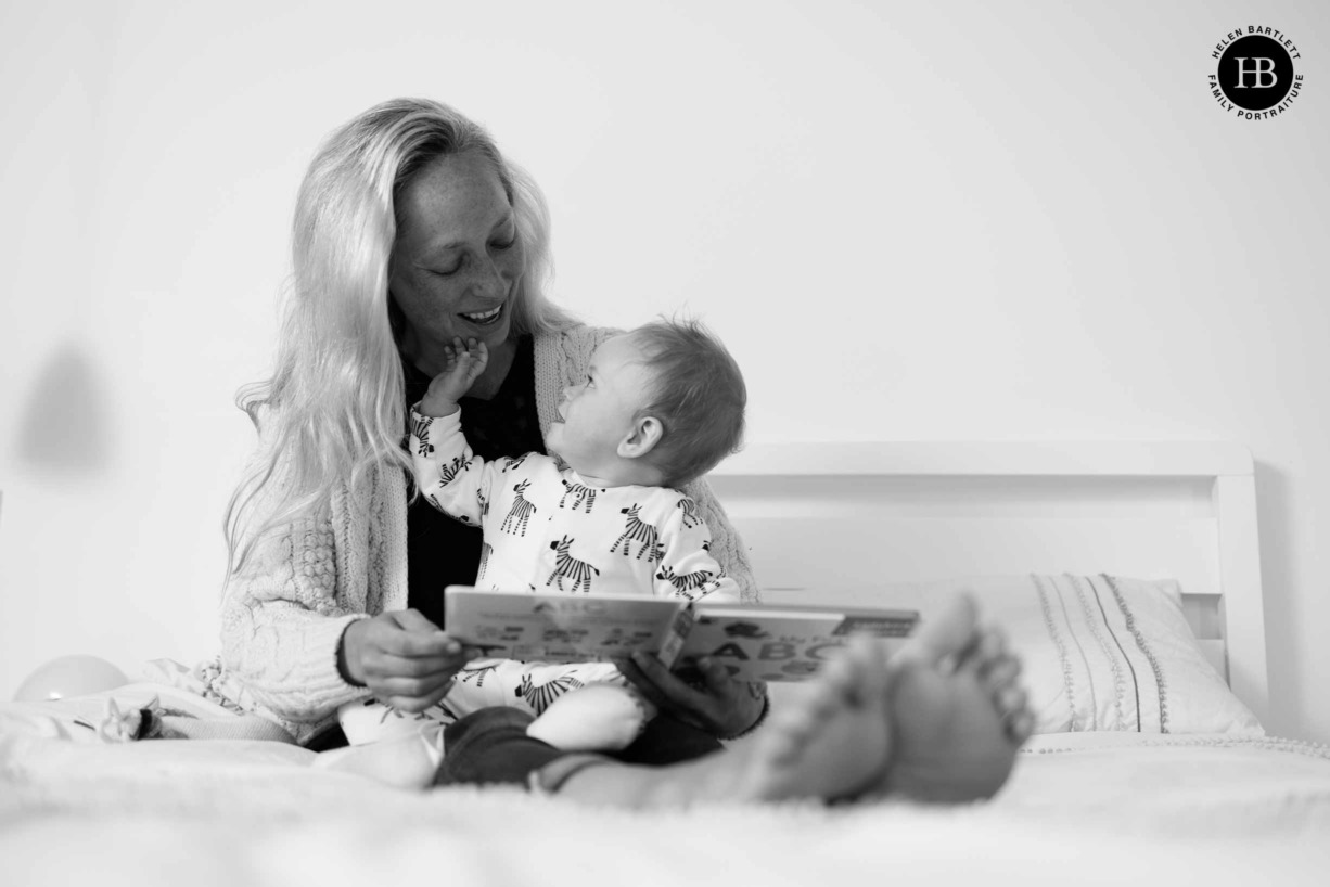 mum and baby sit on the bed reading a story, baby is looking up and touching his mums chin as she looks down. Taken as part of a baby photo shoot in ealing W5