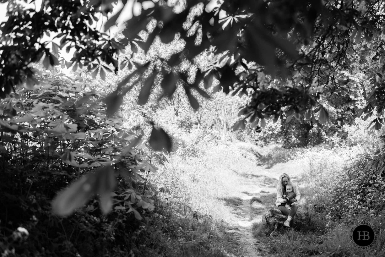 Mum shows baby a flower while sat on a log in the bottom corner of a beautiful wide angle photograph. Foliage is in the foreground creating a tunnel.