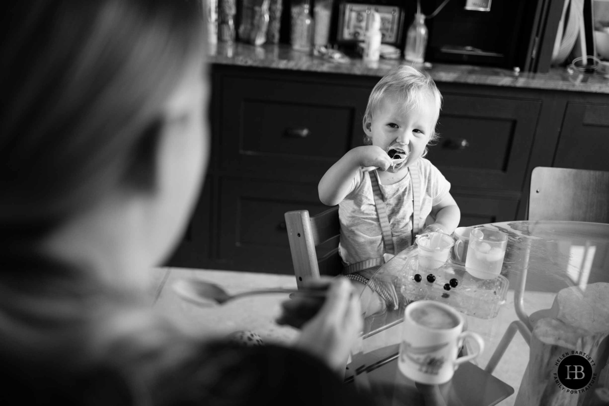 toddler spoons blueberries and yoghurt into their mouth at the breakfast table