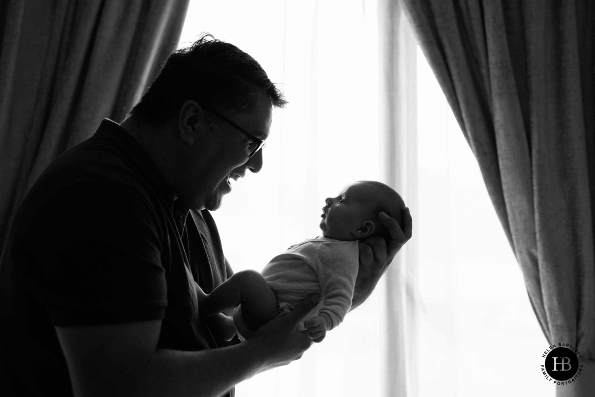 dad-smiles-newborn-baby-held-in-front-of-window